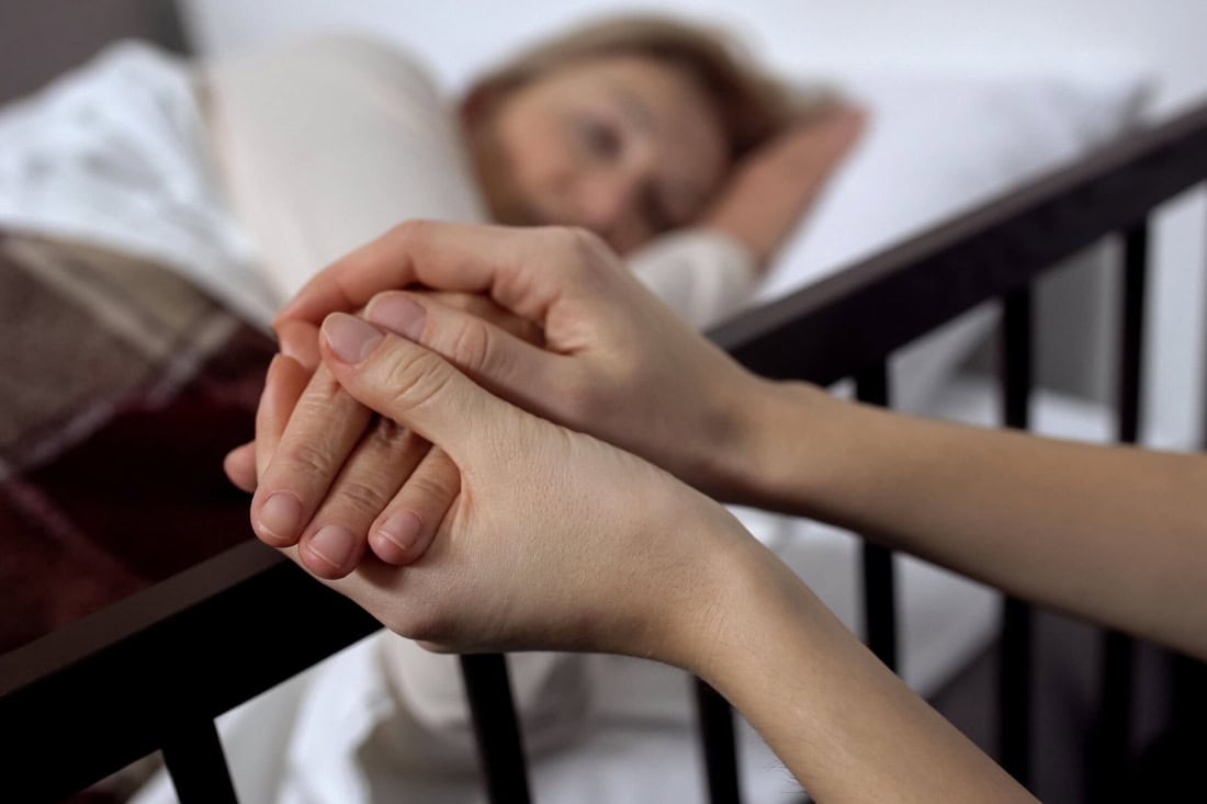 Handen vasthouden van een zieke persoon in bed, symboliseert zorg en steun rondom het levenseinde.